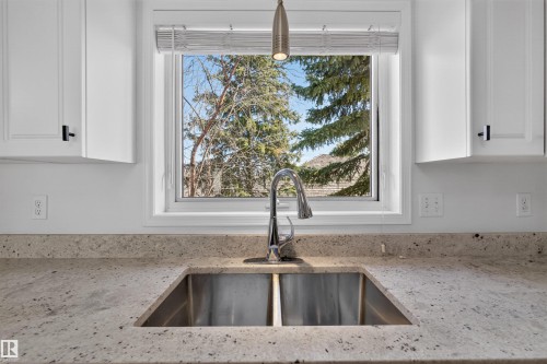 Kitchen sink featuring a dual-basin stainless steel sink, chrome gooseneck faucet, light-toned speckled countertops, white shaker-style cabinetry with matte black hardware, and a window with white blinds - 6 1237 Carter Crest Road, Edmonton, AB 