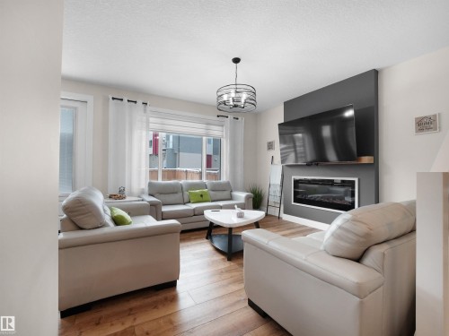 Living area featuring wood-finish flooring, a contemporary ceiling fixture, large windows with white blinds, and an electric fireplace with a dark accent wall - 20827 130 Avenue Nw, Edmonton, AB - Indoor Photo Showing Living Room With Fireplace