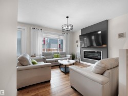 Living area featuring wood-finish flooring, a contemporary ceiling fixture, large windows with white blinds, and an electric fireplace with a dark accent wall - 