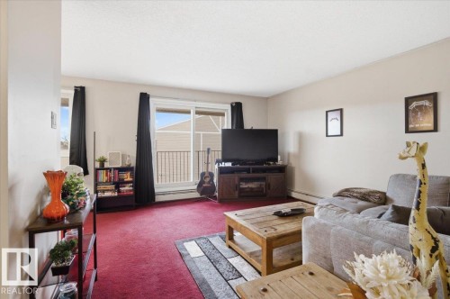Bright living area featuring red carpeting, light-toned walls, and a sliding glass door providing access to an outdoor balcony with a black metal railing - 6 3803 76 Street, Edmonton, AB - Indoor Photo Showing Living Room