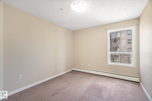Neutral-toned room featuring a single window, baseboard heating, a ceiling-mounted light fixture, and textured carpeting - 203 155 Edwards Drive, Edmonton, AB - Indoor Photo Showing Other Room