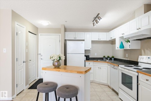 Kitchen featuring white cabinetry, dark countertops, white appliances, and a central island with a wood-finish top - 203 155 Edwards Drive, Edmonton, AB - Indoor Photo Showing Kitchen