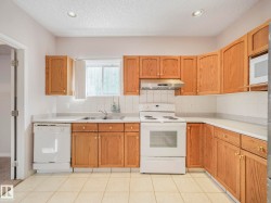 Light-filled kitchen featuring extensive wood-finish cabinetry, white tile flooring, white countertop surfaces, and a white tile backsplash - 