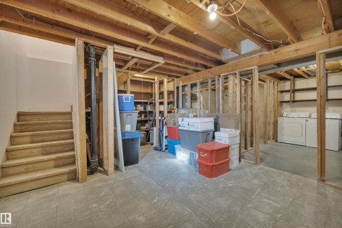 The unfinished basement features exposed wood framing and ceiling joists, a staircase with wooden treads, and a utility area with two washing machines and shelving - 585 Knottwood Road W, Edmonton, AB - Indoor Photo Showing Basement