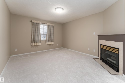 Spacious room featuring light-colored carpeting, a window with curtains, and a fireplace with a tiled surround - 3 9703 174 Street, Edmonton, AB - Indoor With Fireplace