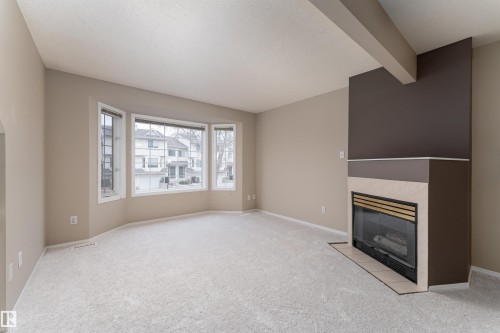 Living area with light-colored carpeting, a bay window, and a corner fireplace featuring a dark brown and light-colored tile surround - 3 9703 174 Street, Edmonton, AB - Indoor With Fireplace