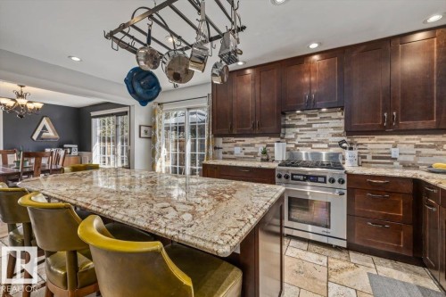 Kitchen featuring a large central island with a stone countertop, dark wood-finish cabinetry, stainless steel range, mosaic tile backsplash, and stone tile flooring - 6003 176 Street, Edmonton, AB - Indoor Photo Showing Kitchen With Upgraded Kitchen