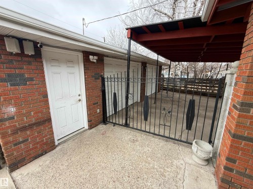 Brick exterior with a white panel door, featuring a concrete patio area, black wrought iron fencing, and a covered carport with a red-stained wooden frame - 11948 132 Street, Edmonton, AB - Outdoor With Exterior