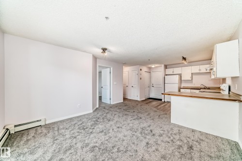 Open-concept living space featuring gray textured carpeting, neutral wall finishes, and a kitchen with white cabinetry and a built-in breakfast bar - 218 10535 122 Street, Edmonton, AB - Indoor