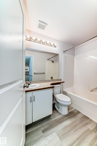 Contemporary bathroom featuring wood-finish flooring, a white vanity with integrated sink, a large wall-mounted mirror, a modern toilet, and a white bathtub with shower - 218 10535 122 Street, Edmonton, AB - Indoor Photo Showing Bathroom