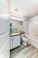 Contemporary bathroom featuring wood-finish flooring, a white vanity with integrated sink, a large wall-mounted mirror, a modern toilet, and a white bathtub with shower - 