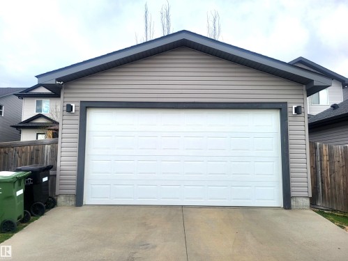 Detached garage featuring tan horizontal siding, a white panel garage door, dark gray trim, and dark gray shingles - 144 Kirpatrick Way, Leduc, AB - Outdoor With Exterior