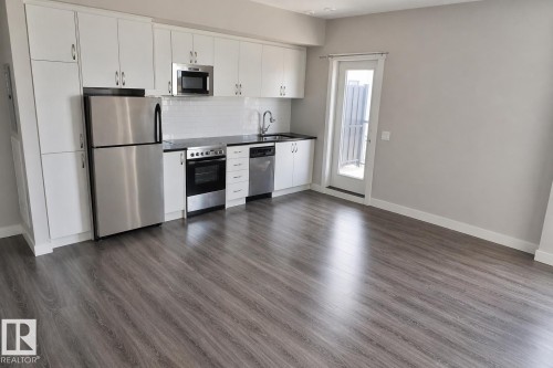 Contemporary kitchen featuring white cabinetry, stainless steel appliances, dark countertops, a white subway tile backsplash, and wood-finish flooring - 81 446 Allard Blvd, Edmonton, AB - Indoor Photo Showing Kitchen