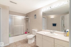 The bathroom features a vanity with a white countertop and an integrated sink, a large mirror with overhead lighting, a toilet, and a bathtub with a sliding glass door enclosure - 