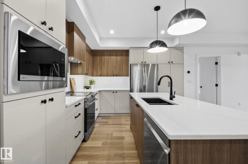 Modern kitchen featuring an extended island with a black sink and matte black faucet, stainless steel appliances, wood-finish flooring, recessed lighting, and elegant pendant fixtures - 202 7459 May Common, Edmonton, AB - Indoor Photo Showing Kitchen With Double Sink With Upgraded Kitchen
