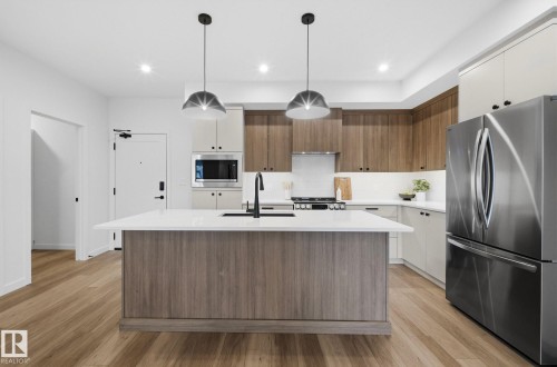 Kitchen featuring a central island with a solid surface countertop and undermount sink, dual pendant lighting, wood-finish flooring, two-tone cabinetry, and stainless steel appliances - 202 7459 May Common, Edmonton, AB - Indoor Photo Showing Kitchen