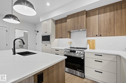 Contemporary kitchen featuring a large island with an undermount sink and matte black faucet, stainless steel appliances, wood-finish cabinetry, white countertops, and a white tile backsplash - 202 7459 May Common, Edmonton, AB - Indoor Photo Showing Kitchen With Double Sink