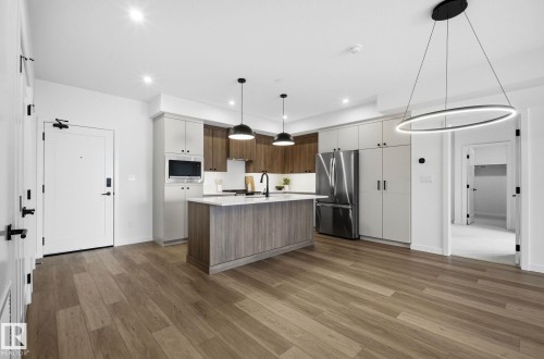 Open-concept living space featuring wood-finish flooring, a contemporary kitchen island with a light-toned countertop, stainless steel appliances, and sleek modern light fixtures - 202 7459 May Common, Edmonton, AB - Indoor Photo Showing Kitchen
