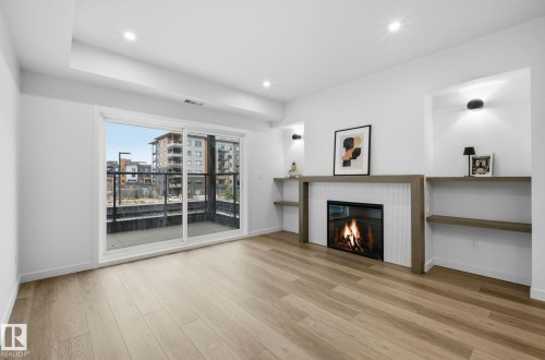 Living area featuring wood-finish flooring, a contemporary fireplace with a white tile surround, built-in shelving, recessed lighting, and sliding glass doors opening to an outdoor balcony with a glass railing - 202 7459 May Common, Edmonton, AB - Indoor Photo Showing Living Room With Fireplace