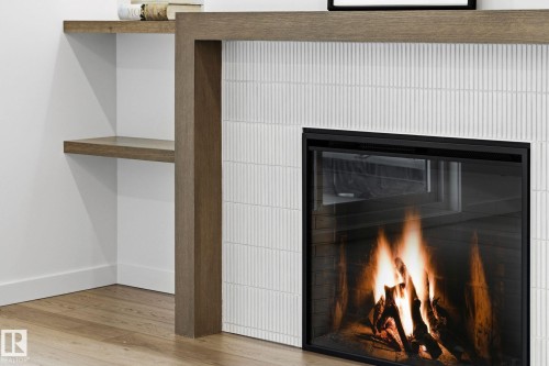 Modern fireplace featuring a textured tile surround, wood-finish mantel, and built-in shelving - 202 7459 May Common, Edmonton, AB - Indoor Photo Showing Other Room With Fireplace