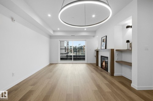Spacious room featuring wood-finish flooring, a contemporary linear fireplace with a wood mantel, and built-in shelving - 202 7459 May Common, Edmonton, AB - Indoor Photo Showing Other Room With Fireplace