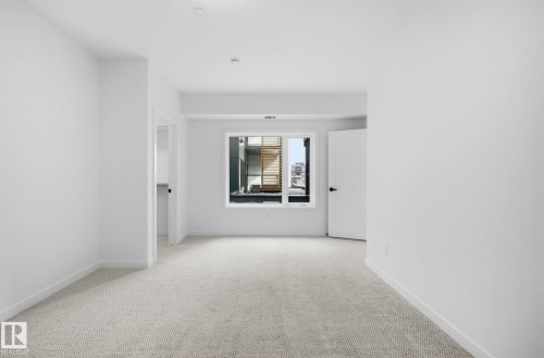 Neutral-toned carpet flooring in a spacious room with a large window, white trim, and a white interior door with black hardware - 202 7459 May Common, Edmonton, AB - Indoor Photo Showing Other Room