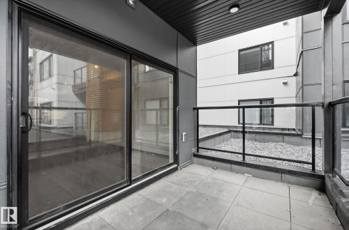 Private patio featuring large format tile flooring, black framed glass sliding doors, and a glass panel railing - 202 7459 May Common, Edmonton, AB - Outdoor With Balcony With Exterior
