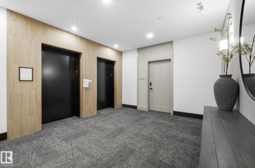 Contemporary hallway featuring two black elevator doors, textured wood-finish wall paneling, and recessed lighting - 202 7459 May Common, Edmonton, AB - Indoor