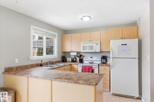 Kitchen featuring wood-finish cabinetry, a double basin stainless steel sink, a window with blinds, and white appliances including a refrigerator, range, and microwave - 70 Stirling Road, Edmonton, AB - Indoor Photo Showing Kitchen With Double Sink