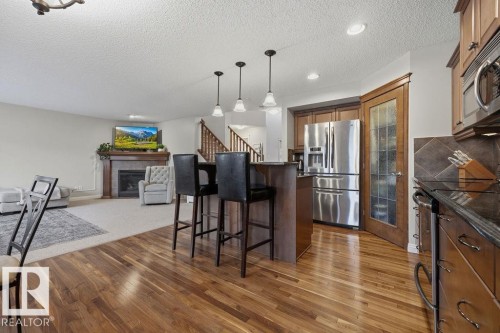 Spacious open-concept interior featuring wood-finish flooring, a kitchen island with pendant lighting, stainless steel refrigerator, dark wood cabinetry, and a fireplace with a stone surround - 1613 Kerr Road, Edmonton, AB - Indoor Photo Showing Kitchen With Stainless Steel Kitchen With Upgraded Kitchen