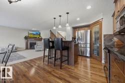 Spacious open-concept interior featuring wood-finish flooring, a kitchen island with pendant lighting, stainless steel refrigerator, dark wood cabinetry, and a fireplace with a stone surround - 