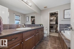 Bathroom featuring a single vanity with wood cabinetry, a large mirror, a built-in tub with tile surround, an open closet, and tile flooring - 