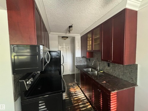 Galley kitchen featuring rich wood-finish cabinetry with crown molding, dark stone countertops, a double basin stainless steel sink with gooseneck faucet, and a stone tile backsplash - 111 10225 117 Street, Edmonton, AB 