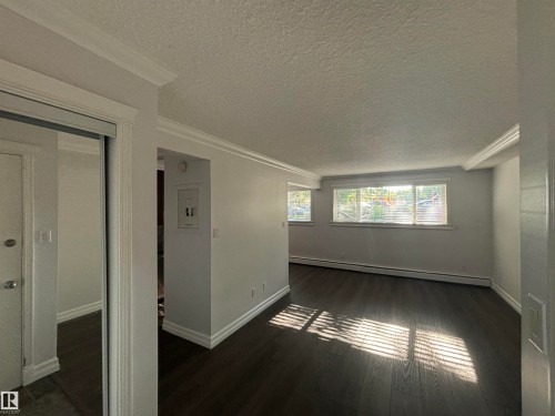 Spacious room featuring dark wood-finish flooring, multiple windows, white baseboards, and white crown molding - 111 10225 117 Street, Edmonton, AB 