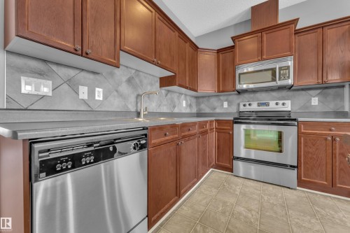 Kitchen featuring wood cabinetry with knob hardware, stainless steel appliances including a built-in microwave, a stainless steel sink with a high-arch faucet, and a patterned tile backsplash - 110 13825 155, Edmonton, AB - Indoor Photo Showing Kitchen