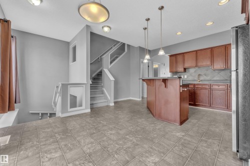 Open-concept kitchen and entryway featuring a central island with pendant lighting, wood cabinetry, tile flooring, and a multi-level staircase with white railings - 110 13825 155, Edmonton, AB - Indoor Photo Showing Kitchen