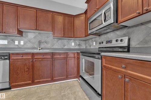 Kitchen featuring extensive wood cabinetry, stainless steel appliances, a tile backsplash, composite countertops, and tile flooring - 110 13825 155, Edmonton, AB - Indoor Photo Showing Kitchen