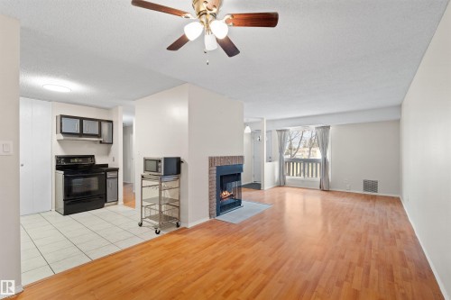 Open concept living area featuring wood-finish flooring, a brick fireplace with a stone hearth, a ceiling fan with integrated lighting, and a kitchen area with tile flooring and a black electric range - 1116 Saddleback Road, Edmonton, AB - Indoor Photo Showing Living Room With Fireplace