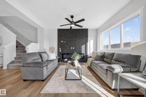 Spacious living area featuring wood-finish flooring, a contemporary fireplace with a vertical slat surround, a ceiling fan, and large windows - 20723 24 Avenue, Edmonton, AB - Indoor Photo Showing Living Room