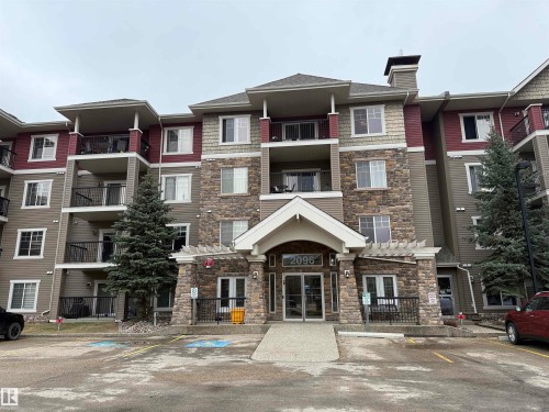 Multi-story residential building featuring a stone and siding exterior, multiple balconies with black railings, a covered entryway with a decorative pergola, and mature evergreen landscaping - 135 2096 Blackmud Creek Drive, Edmonton, AB - Outdoor With Facade