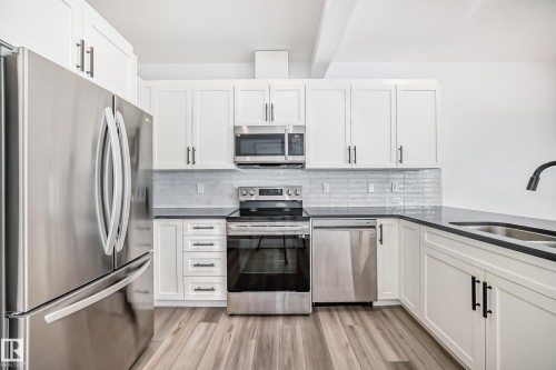 Modern kitchen with white cabinetry and dark hardware, stainless steel appliances, dark countertops, and wood-finish flooring - 38 1910 Collip View, Edmonton, AB - Indoor Photo Showing Kitchen