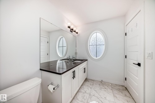 Modern bathroom featuring a double vanity with dark countertops and dual undermount sinks, an oval privacy window, and white panel doors with black hardware - 38 1910 Collip View, Edmonton, AB - Indoor Photo Showing Bathroom