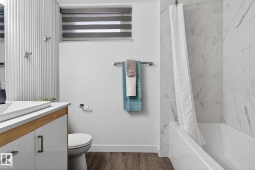 Modern bathroom featuring a soaking tub with large format marble-look tile surround, a built-in vanity with a white countertop and wood-finish accents, and wood-finish flooring - 13332 106 Street, Edmonton, AB - Indoor Photo Showing Bathroom