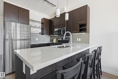 Kitchen featuring a white countertop island with integrated sink, stainless steel appliances, dark wood-finish cabinetry, and a horizontal tile backsplash - 403 10418 81 Avenue, Edmonton, AB - Indoor Photo Showing Kitchen With Double Sink With Upgraded Kitchen