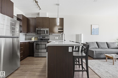 Modern kitchen featuring dark wood-finish cabinetry, stainless steel appliances, light-toned countertops, and a full-height tile backsplash - 403 10418 81 Avenue, Edmonton, AB - Indoor Photo Showing Kitchen With Upgraded Kitchen