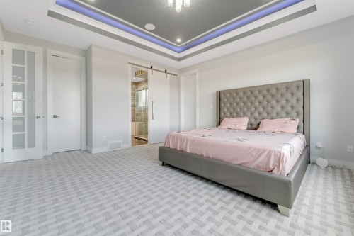 Spacious room featuring patterned carpet flooring, a tray ceiling with accent lighting, and a barn door entry to a tiled bathroom with a glass shower enclosure - 3736 1 Avenue Sw, Edmonton, AB - Indoor Photo Showing Bedroom