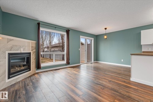 Spacious interior featuring wood-finish flooring, a corner fireplace with a tiled surround, a large picture window, and a glass sliding door - 5407 204 Street, Edmonton, AB - Indoor Photo Showing Living Room With Fireplace