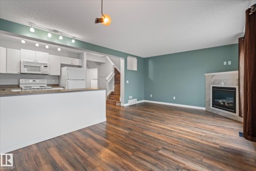 Open-concept living area featuring wood-finish flooring, a contemporary fireplace with a tiled surround, a kitchen bar, white cabinetry, and a full suite of white appliances - 5407 204 Street, Edmonton, AB - Indoor Photo Showing Kitchen With Fireplace
