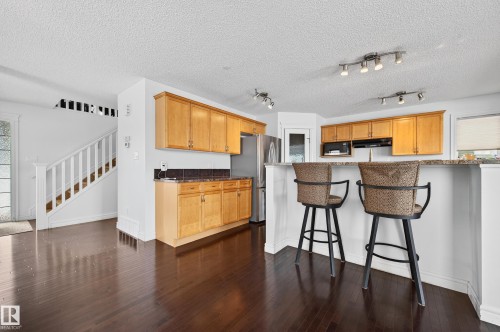 Kitchen area featuring wood-finish flooring, light wood cabinetry, a stainless steel refrigerator, and a stone-finish countertop with a breakfast bar - 8477 Sloane Crescent, Edmonton, AB - Indoor Photo Showing Kitchen