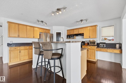 Spacious kitchen featuring warm wood cabinetry, dark wood-finish flooring, a central island with countertop seating, stainless steel appliances, and track lighting fixtures - 8477 Sloane Crescent, Edmonton, AB - Indoor Photo Showing Kitchen